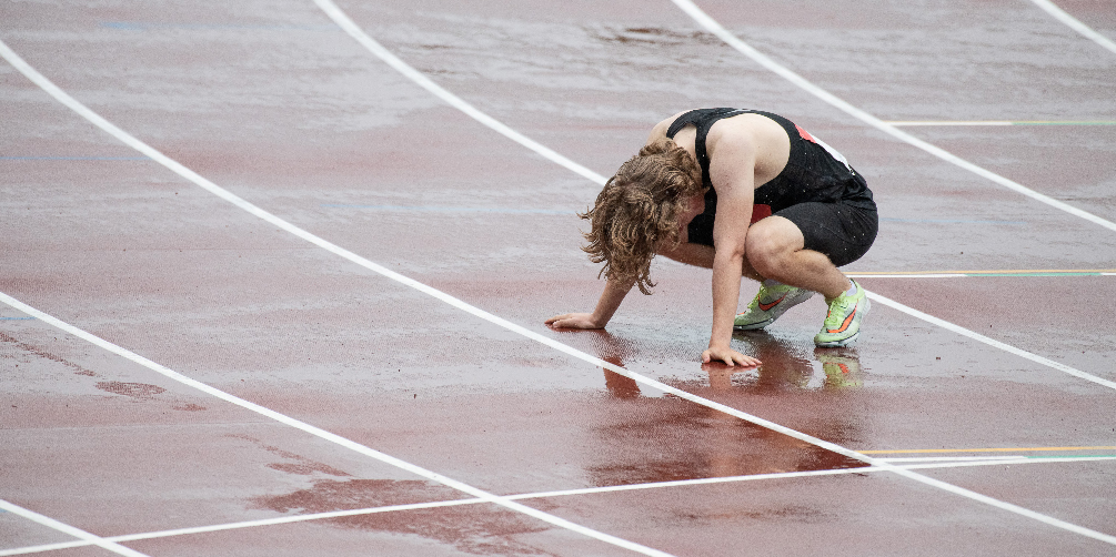 Juoksun jälkeinen uupunut juoksija kyykyssä nojaten käsillään maahan sateisella urheilukentällä. Taustalla näkyy pelkkää urheilukentän punaista pintaa ja juoksuradan viivoja.