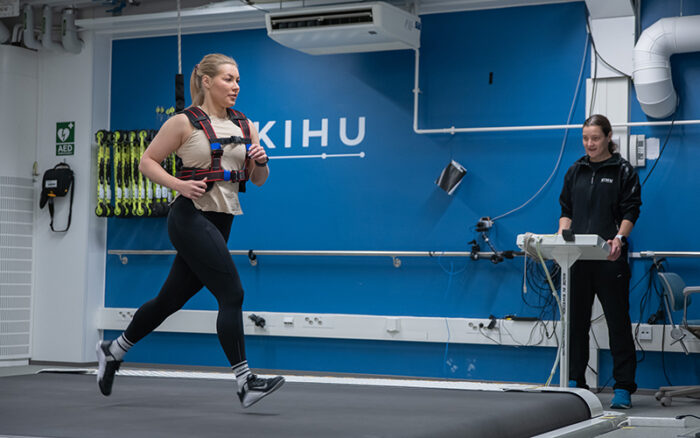 Naisurheilija juoksee juoksumatolla testaajan kannustaessa taustalla ohjauspaneelin ääressä. Juoksumaton takana sininen seinä, jolla roikkuu rullasuksia ja näkyy KIHU-logo.