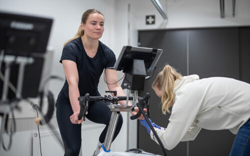 Mustapaitainen nainen polkee kuntopyörää kuntotestissä. Testaaja kumartuneena alas ja ottaa naisen vasemmasta kädestä verinäytettä.